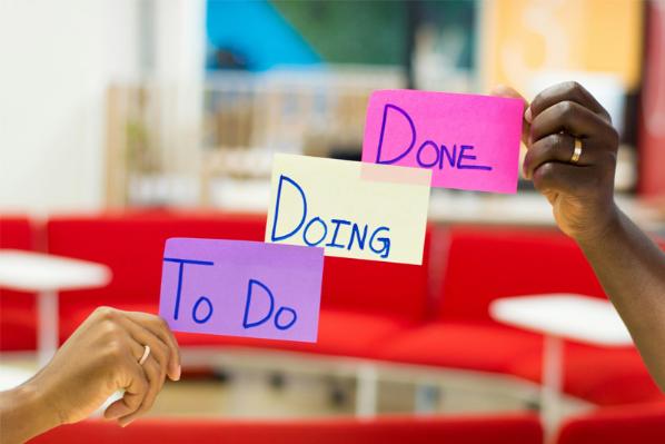 Two people hold three sticky notes reading, To Do, Doing, Done.