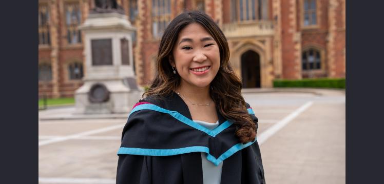 A student pictured in her graduation gown outside university building looking at the camera