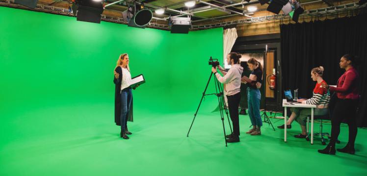 A wide-angle shot of an eclectic group of people working in a film studio, and a woman presenting in front of a green screen