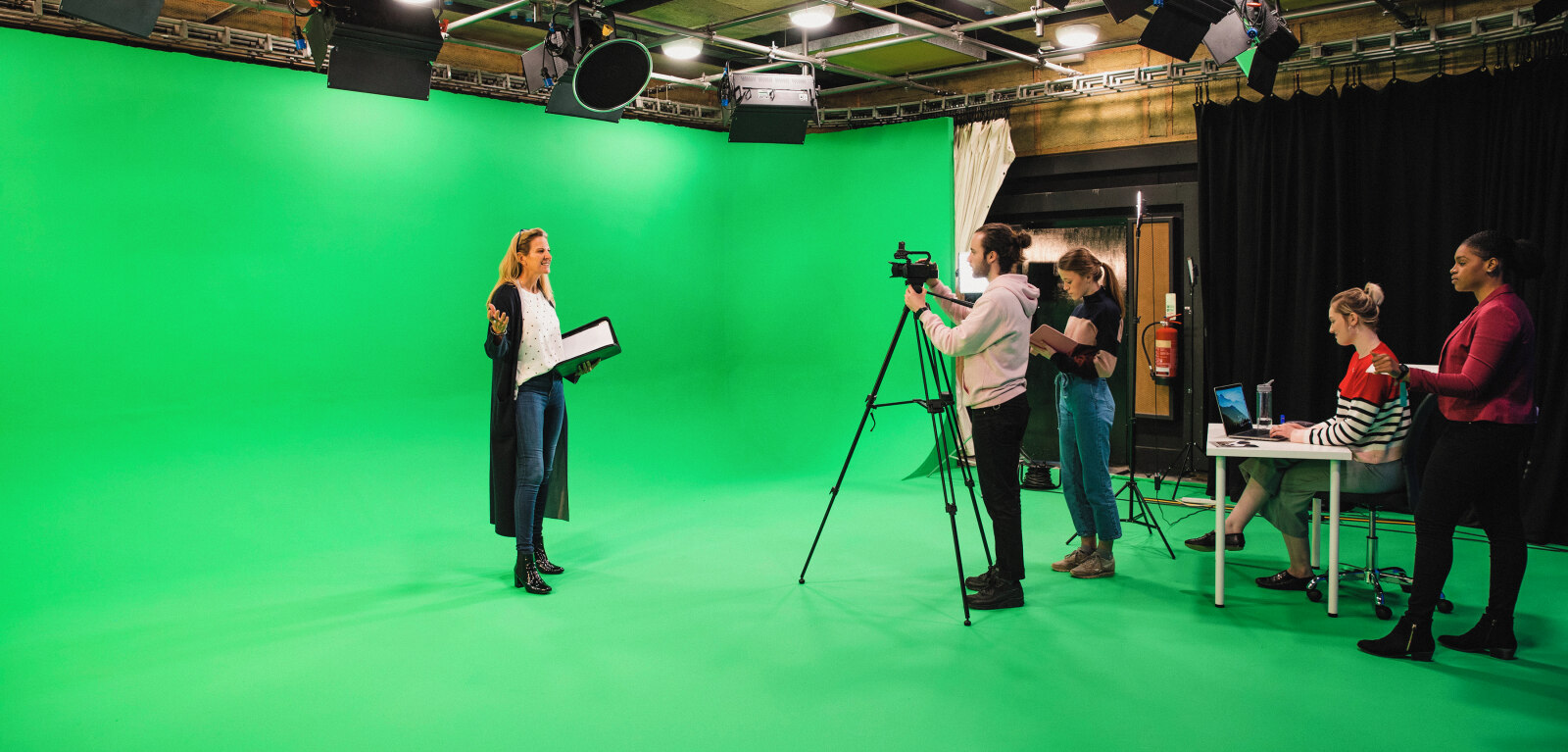 A wide-angle shot of an eclectic group of people working in a film studio, and a woman presenting in front of a green screen