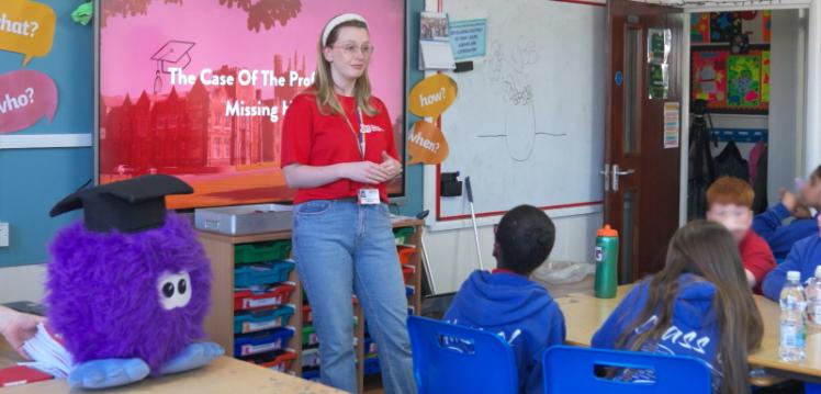 A woman stands at the front of a classroom speaking to young school children. A fluffy purple soft toy sits to the left of the frame.