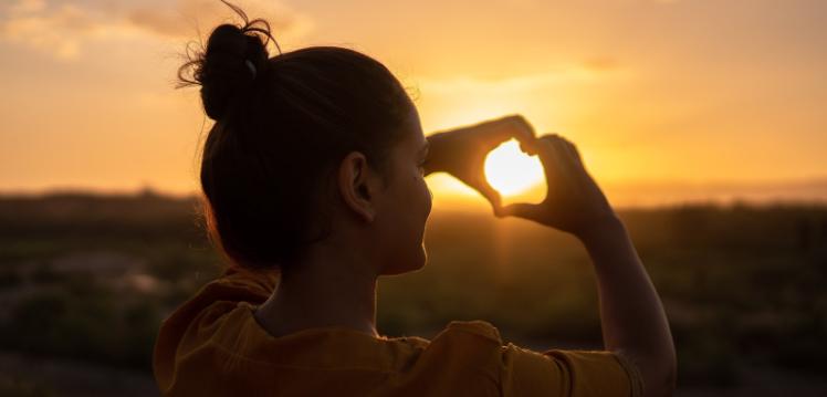 Woman doing heart sign against the sun