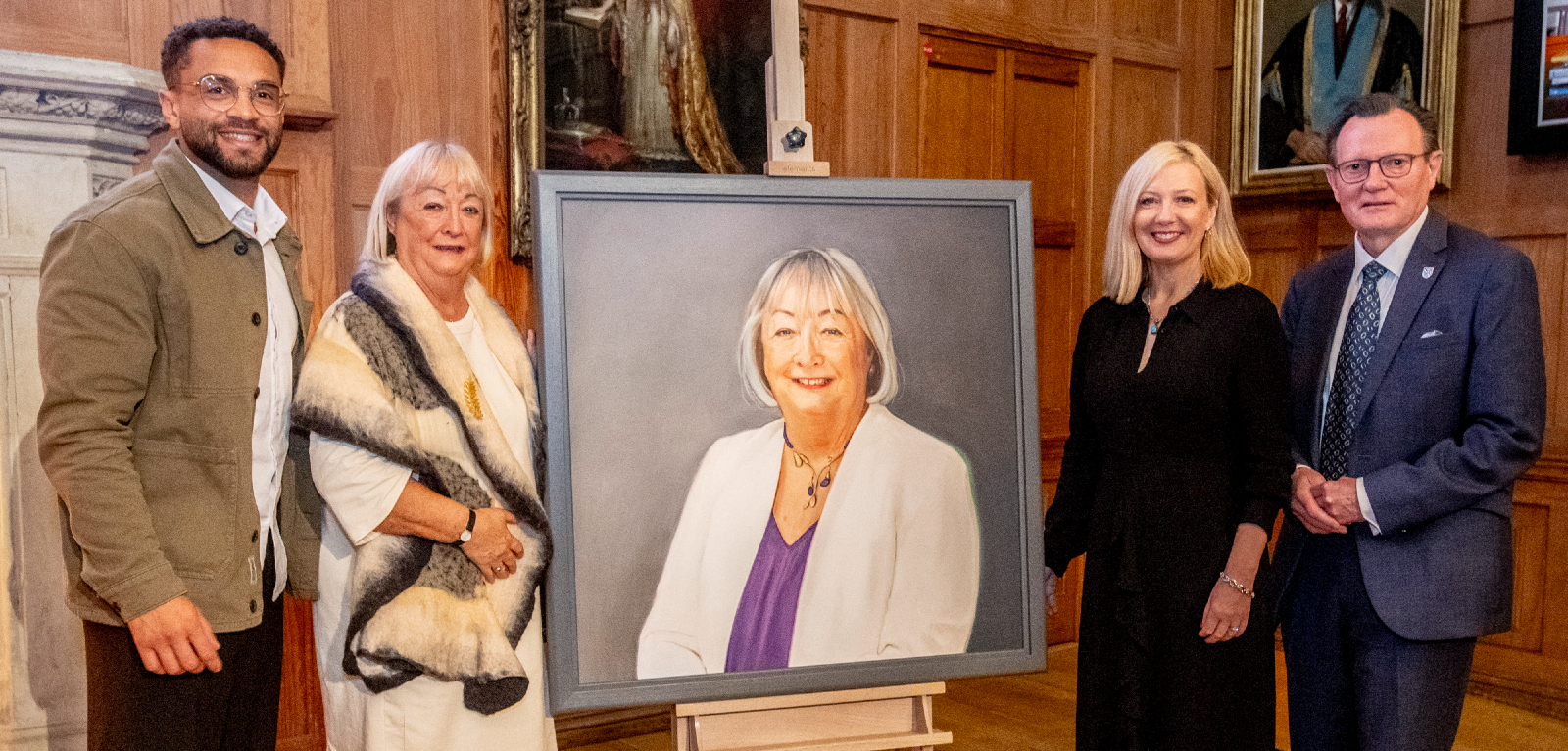 Two men and two women stand either side of a large portrait in a University's grand hall
