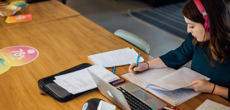 Student writing notes on table while wearing headphones