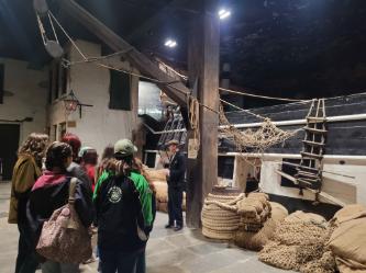 A group of students stood looking at a big ship in a building made to look like a dock with a night’s sky. The photo is taken from behind the students and they are all listening to a man dressed as a shipping clerk complete with a flat cap