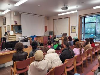 A group of students sat on rows on chairs – they are all facing forward to listen to two older men who are sat at the front in front of a pulled down presentation screen. They are in a library.