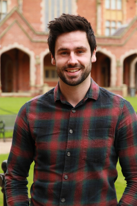 Head and shoulders of Brian Devlin smiling, wearing a red and black shirt, standing in the quad at Queen's