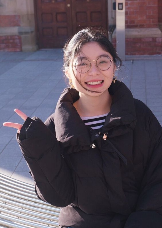 Xi Wang seated on a metal bench in the sunshine on the main Queen's campus. She's smiling, wearing a black padded coat and has her hand raised in greeting.