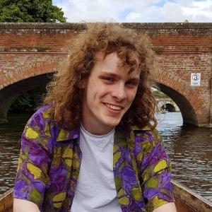 PhD student profile photo, a male in a boat on the water with a bridge behind him