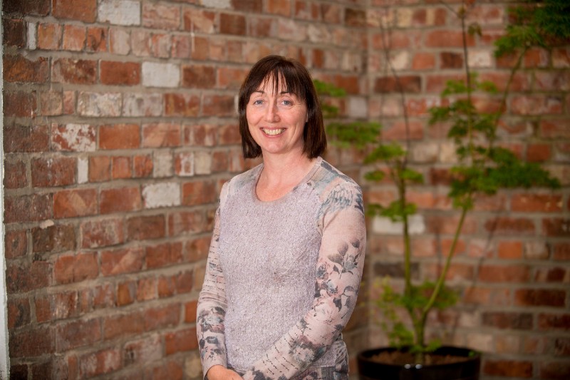 smiling woman with fringe and shoulder length brown hair, against a red brick wall background