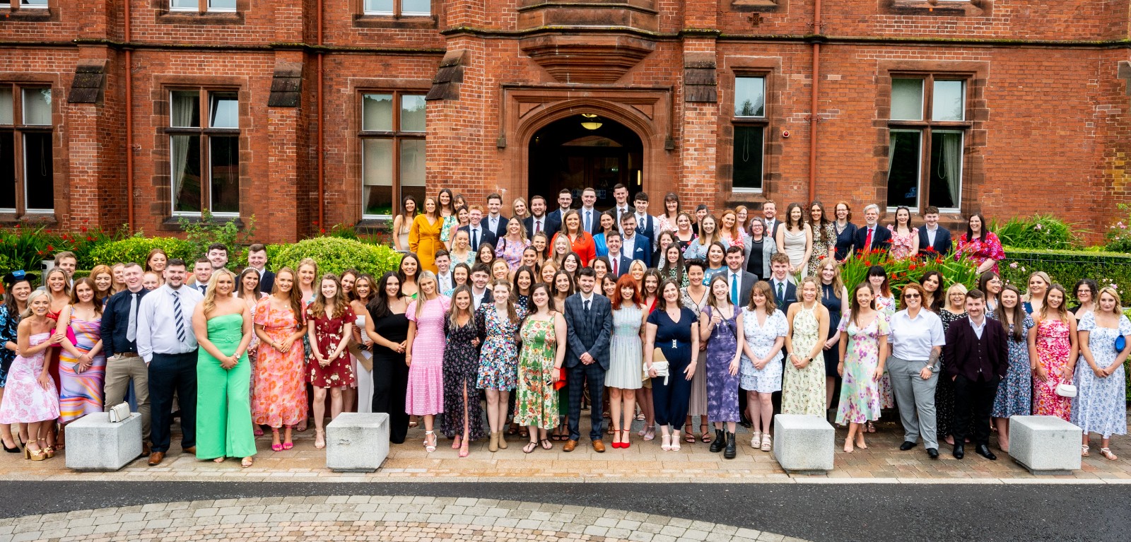 100+ smartly dressed men and women in their early twenties lined up in several rows on the steps leading down from the entrance to a large, red-brick building