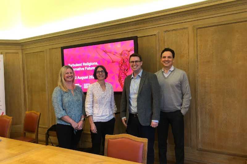 two women and two men standing in front of an oak-panelled wall with a screen showing the conference date and title: Turbulent Religions, Alternative Futures, August 2023