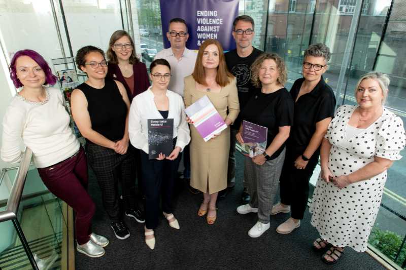 eight women and two men in a row, in front of a banner saying End Violence Against Women and Girls, with three women holding a copy of a report