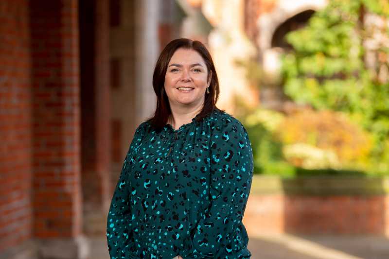 smiling woman with shoulder length dark hair against a background of red brick and green shrubbery