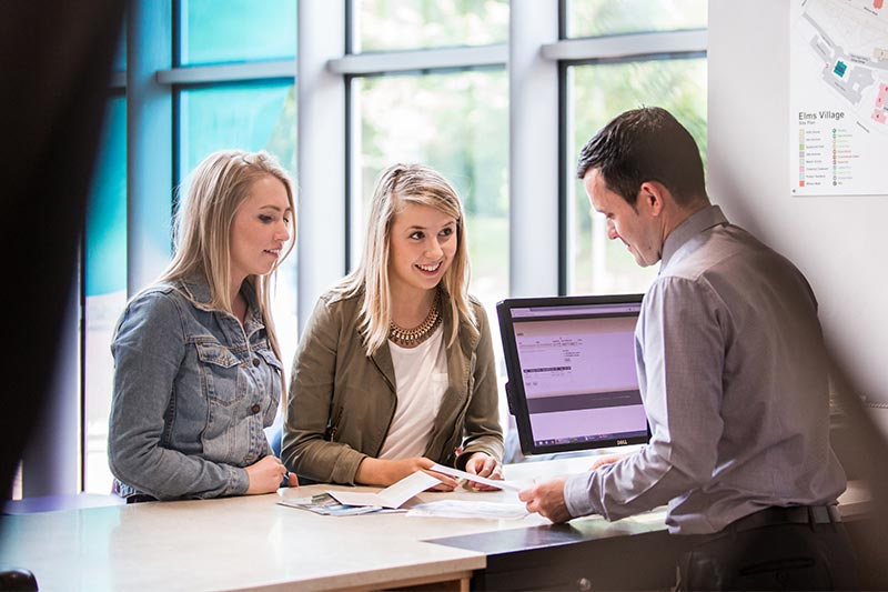 two female students talking to a member of staff at the Elms Village reception desk