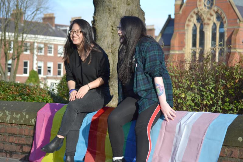 Two female students sit on a wall draped with LGBT flags and Trans rights flags