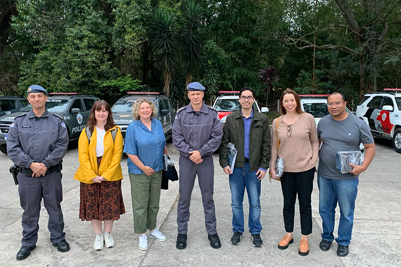 three women and four men, including two policemen, standing in a row in a car park with police vehicles..