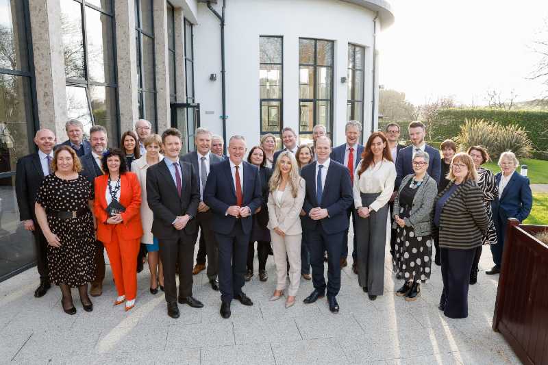 about 25 men and women in smart workwear standing on a patio beside large curved windows