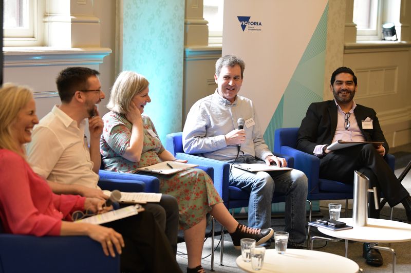 two women and three men seated in a row in a brightly lit room, all smiling, with one man holding a microphone