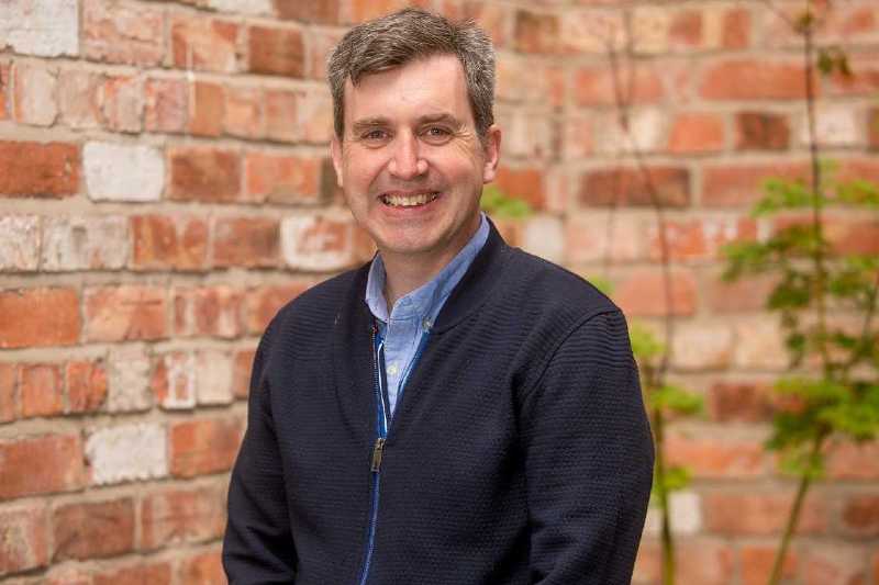 smiling man with thick greying hair, wearing a light blue shirt and havy zip jacket, with a red brick wall behind