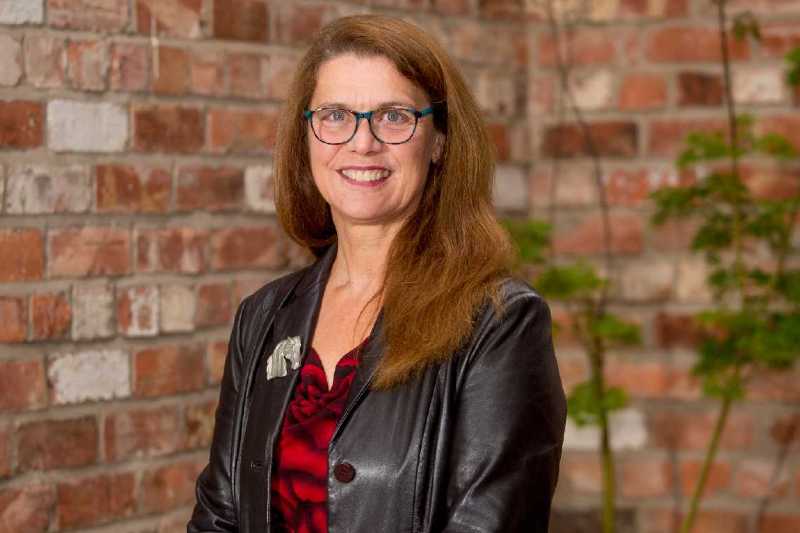 dark haired woman in glasses and a black leather jacket against a red brick background