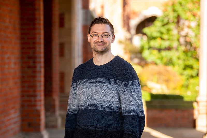 man with short dark hair, dark beard and black rimmed glasses wearing a black and grey jumper and with green foliage behind him