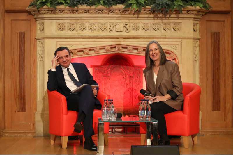 dark haired man in a dark suit and woman with shoulder length hair and brown jacket, both seated in front of a large stone fireplace