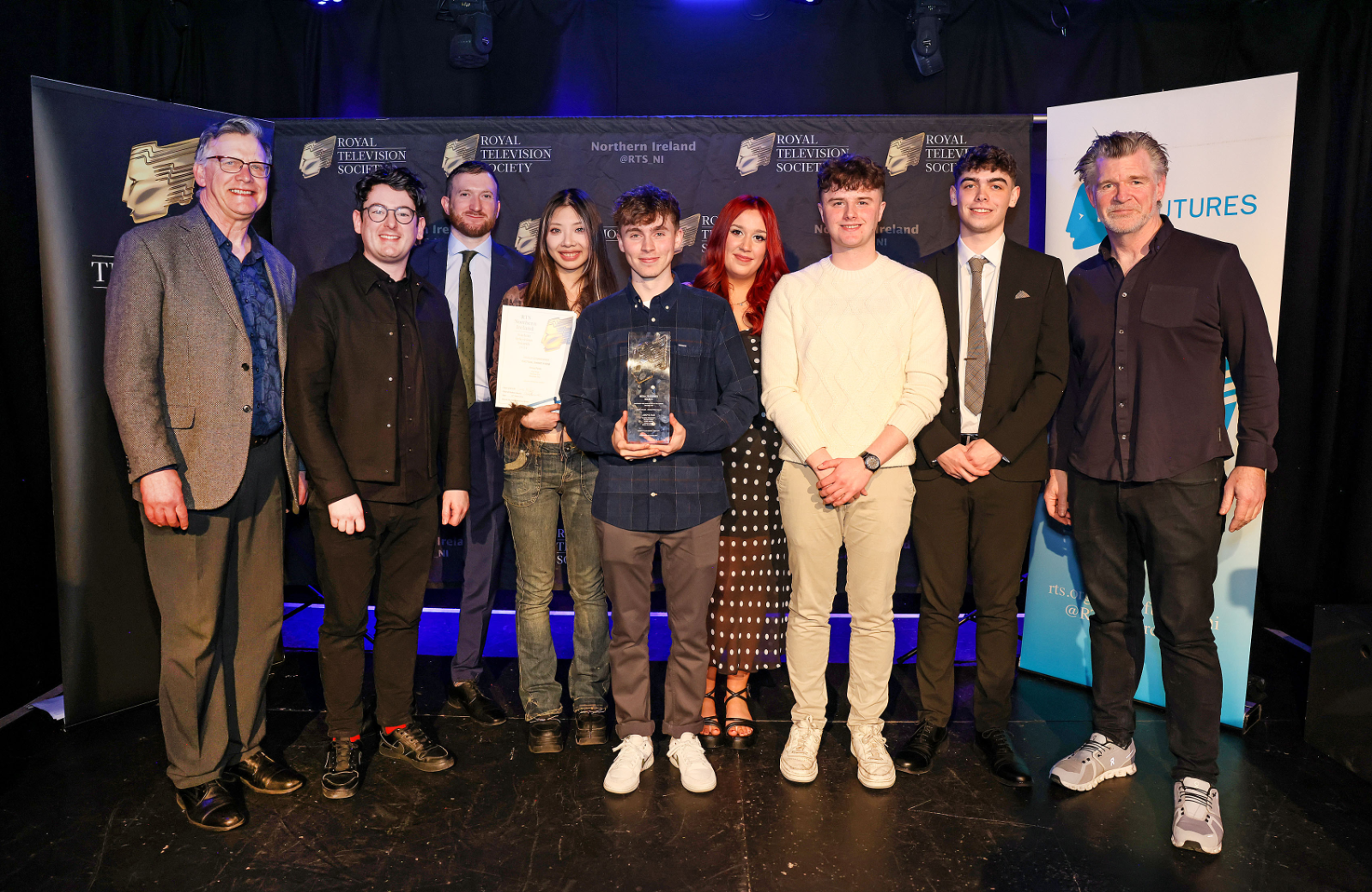 A group of people standing in a semicircle in front of display stands. The person in the centre is holding an award