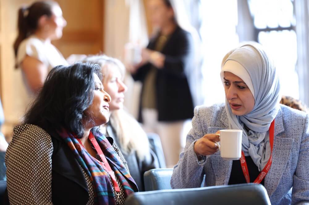 Two staff members engaged in conversation, one holding a coffee cup, with other people interacting in the background