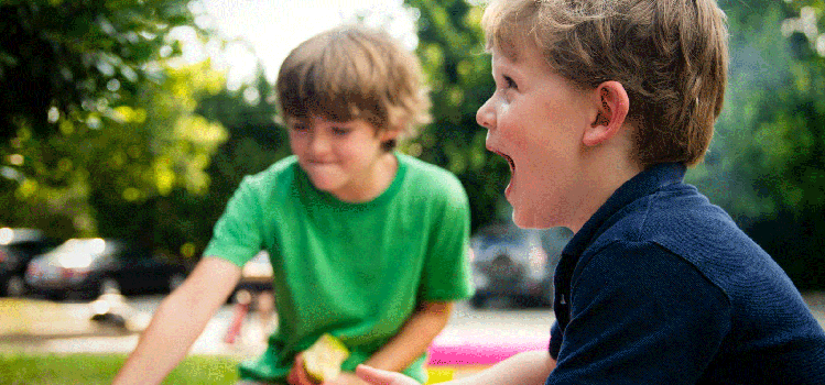 Two young boys sitting outside on a sunny day, one wearing a green shirt and smiling while holding a piece of watermelon, the other in a navy shirt laughing with excitement.