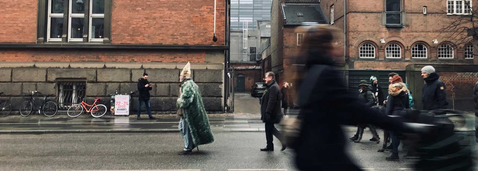 a man wearing a long green coat and a pointed hat leads a group of people walking along the middle of a wet road. A woman on a bicycle is in the foreground going in the opposite direction.