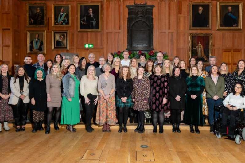 three standing rows of men and women with a wood panelled wall behind