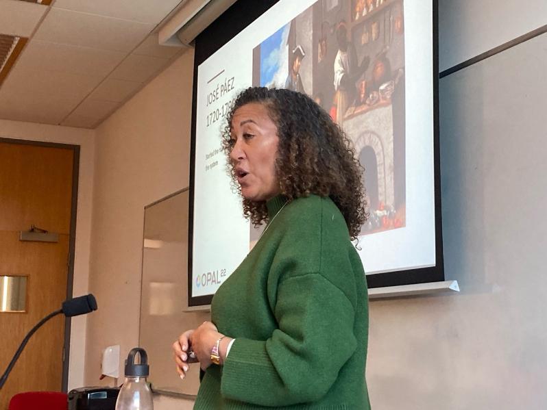 Woman with mid-length, dark, curly hair and wearing a green jumper delivering a presentation