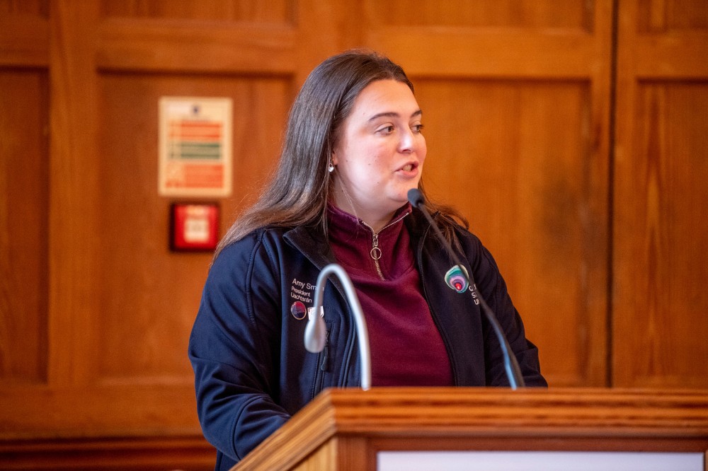young woman wearing navy blue sports top addressing an indoor event from a podium