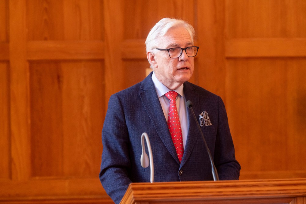 white-haired man in dark blue suit, light shirt and red tie standing at a podium addressing an indoor event