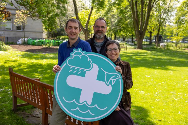 Two men and one woman stand in a garden holding a teal logo sign.