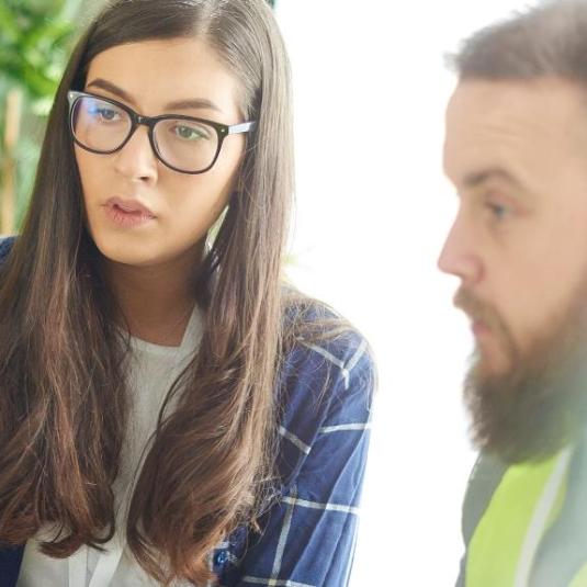 young woman wearing glasses talking with a young bearded man in an office environment