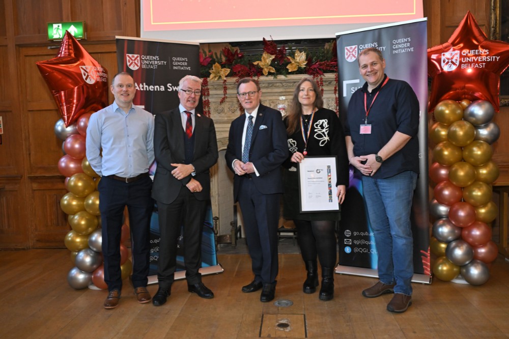 a woman holding a framed award certificate poses for a photo with four men in an old wood-paneled hall with large decorated fireplace and presentation screen in the background, framed by two celebratory balloon pillars