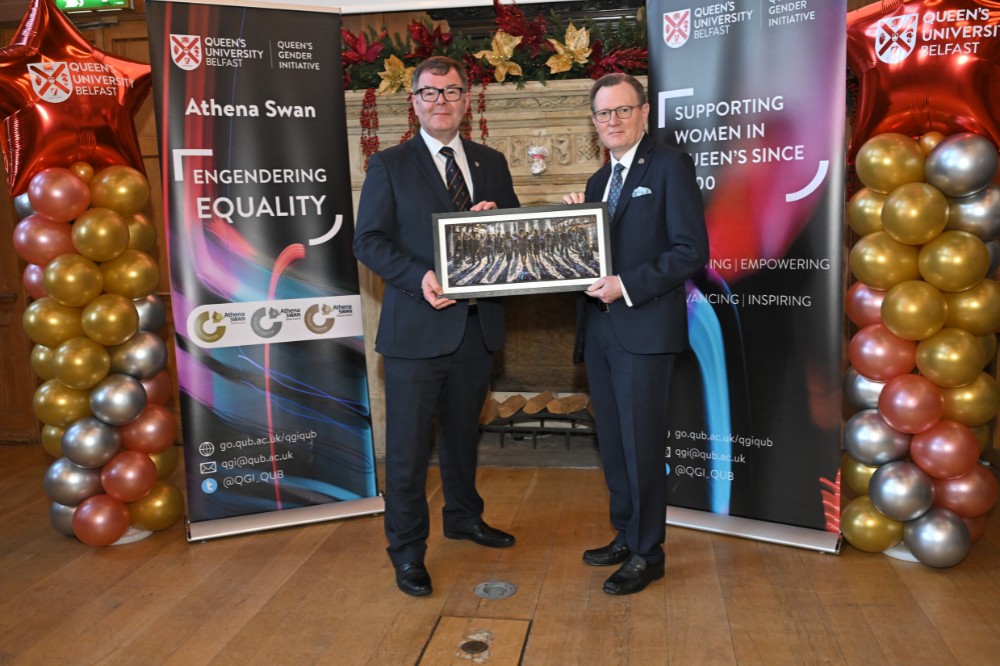two besuited men posing with a framed painting in an old event hall with large fireplace in the background and pop-up stands and balloon pillars framing them