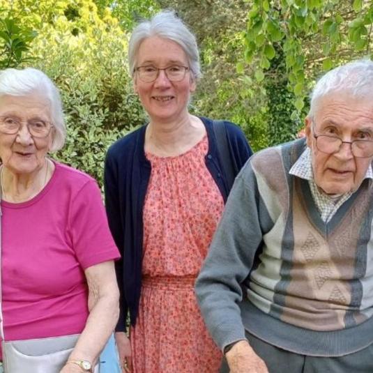 elderly woman and man standing with younger woman in middle with green shrubbery in the background