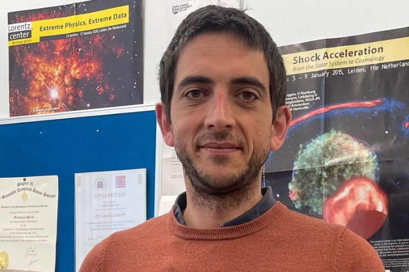 smiling hirsute man standing in front of physics research event posters