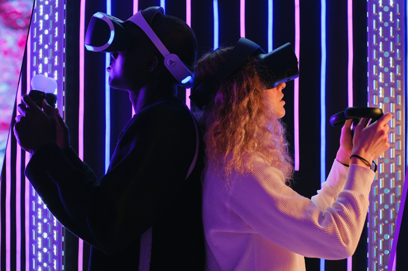 man and woman with their backs to each other in a dim, technological looking indoor space using XR or VAR headsets and controllers