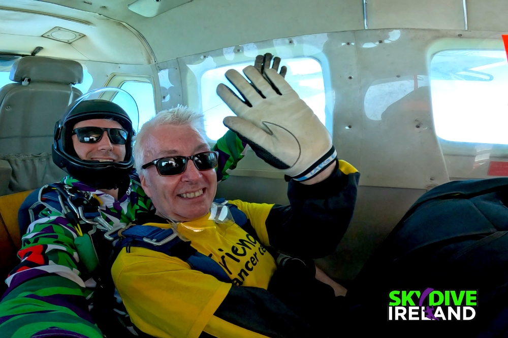 seated man waving to camera inside a plane, in front of instructor, waiting to do a skydive