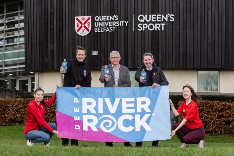 three men standing behind a Deep RiverRock advertising banner, each holding a bottle of water, and two women in red jumpers kneeling on either side of the banner helping to hold it up