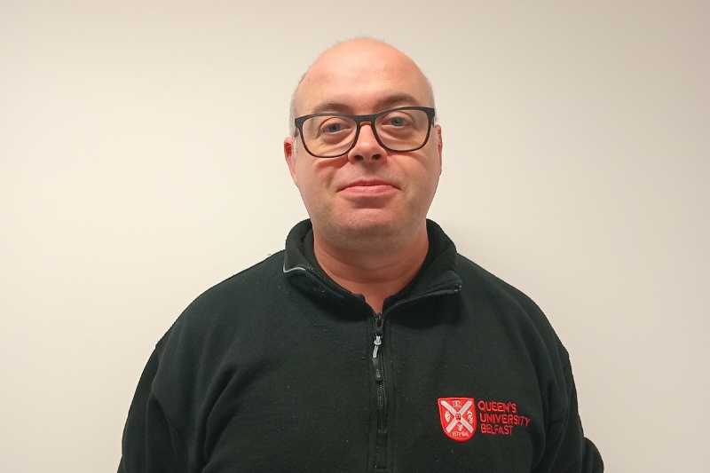 A man with glasses and a neutral expression standing in front of a plain white background. He is wearing a black fleece jacket with the Queen's University Belfast logo on the left side.