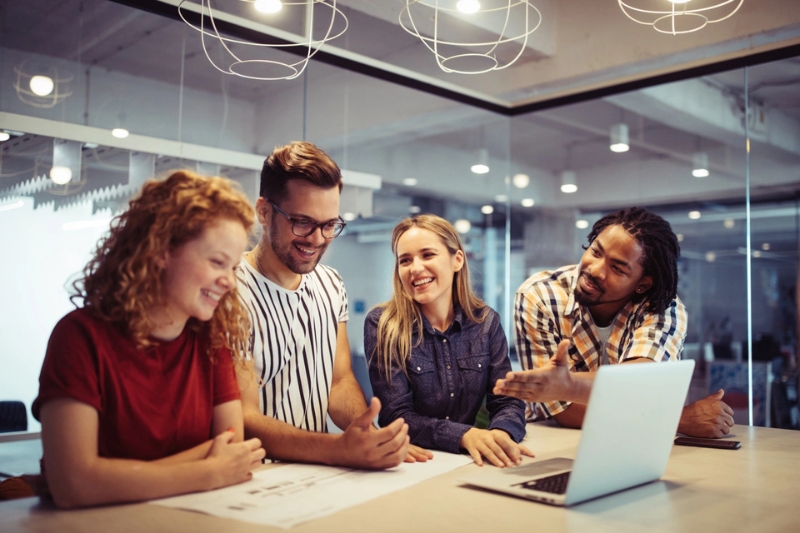 two males and two females smiling and discussing something over a table at a laptop in a group study space