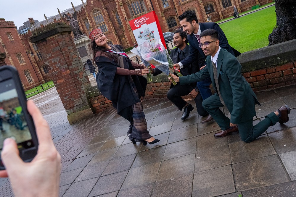 three men jokingly bent on one knee offering bouquets of flowers to a smiling woman in front of the gates of an old redbrick building