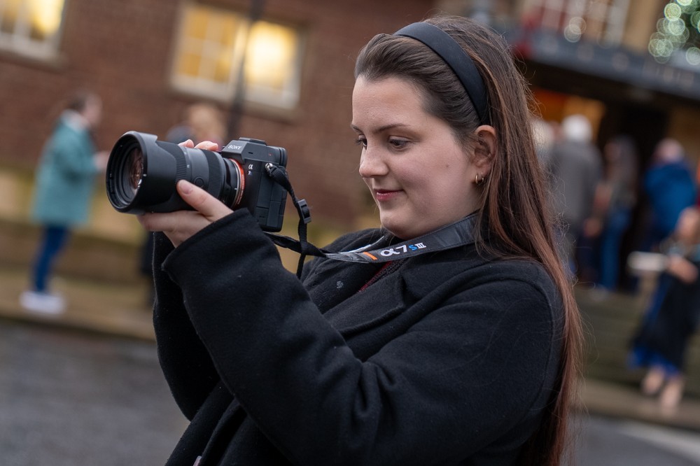 happy looking young woman in black coat taking photos with her camera outdoors
