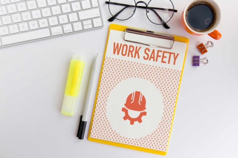 home or office desk with laptop keyboard, pen, highlighter, cup of coffee, folded spectacles, and clipboard holding 'work safety' training sheets